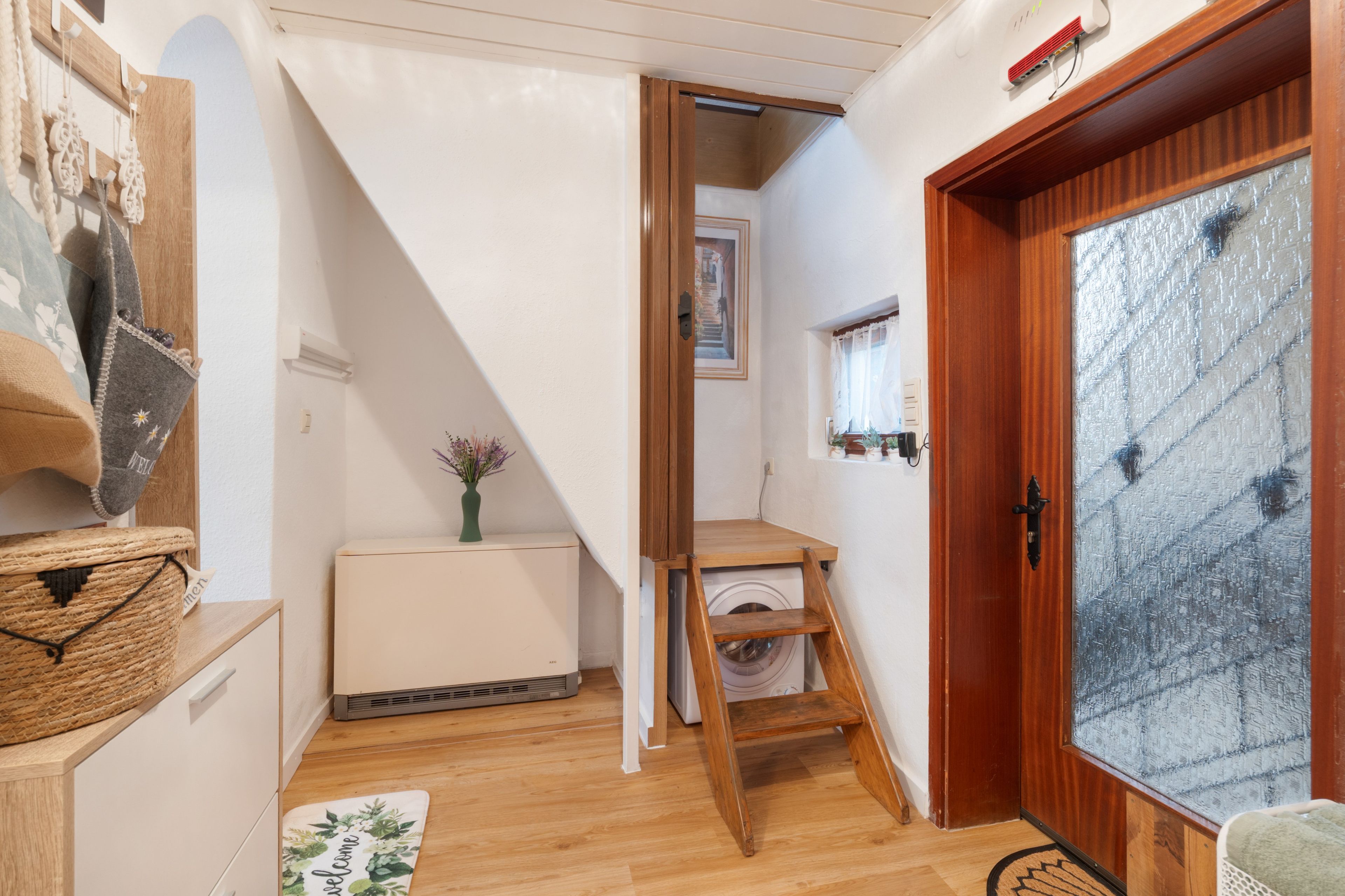 A narrow hallway with a wooden door, a radiator, a small table with a vase, and a welcome mat.