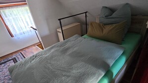 Een enkel bed met een groene matras en een grijze deken in een kamer met een schuine plafond.