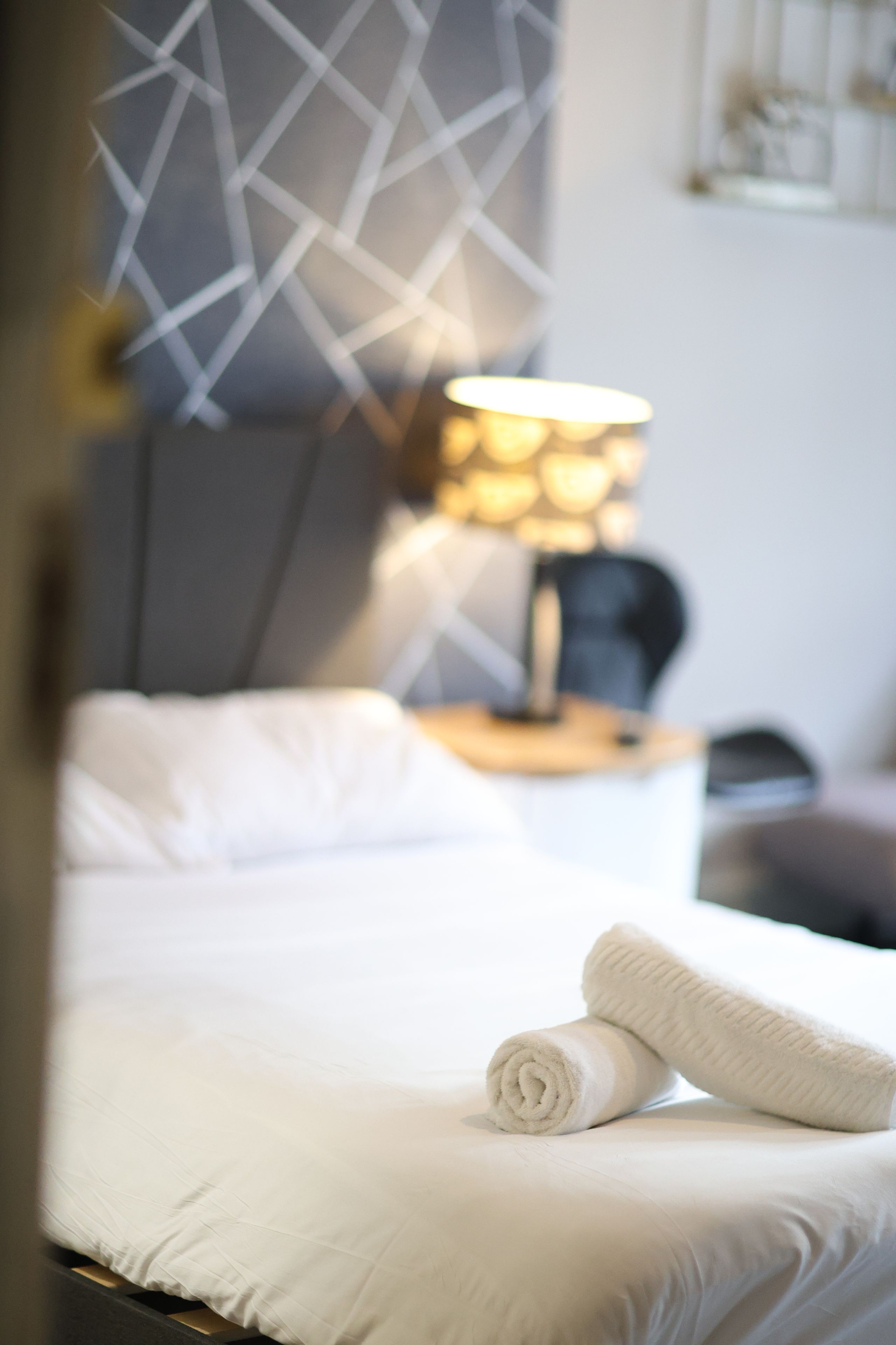 Cama bem arrumada com toalha branca enrolada, abajur e cabeceira geométrica moderna.