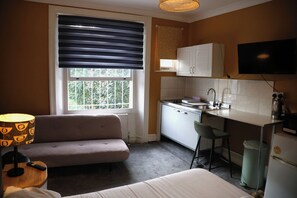 Een compact keukenhoekje met een raam, een bank, een klein tafeltje en een aan de muur bevestigde televisie.
