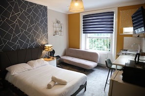 A hotel room with a bed, a sofa, a desk, a lamp, and a window with blinds. - Gold Grey Studio (London)