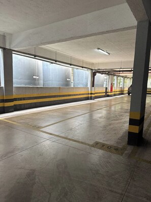 Un estacionamiento subterráneo vacío con pisos de concreto, barreras rayadas de amarillo y negro, y una puerta roja de salida de emergencia.