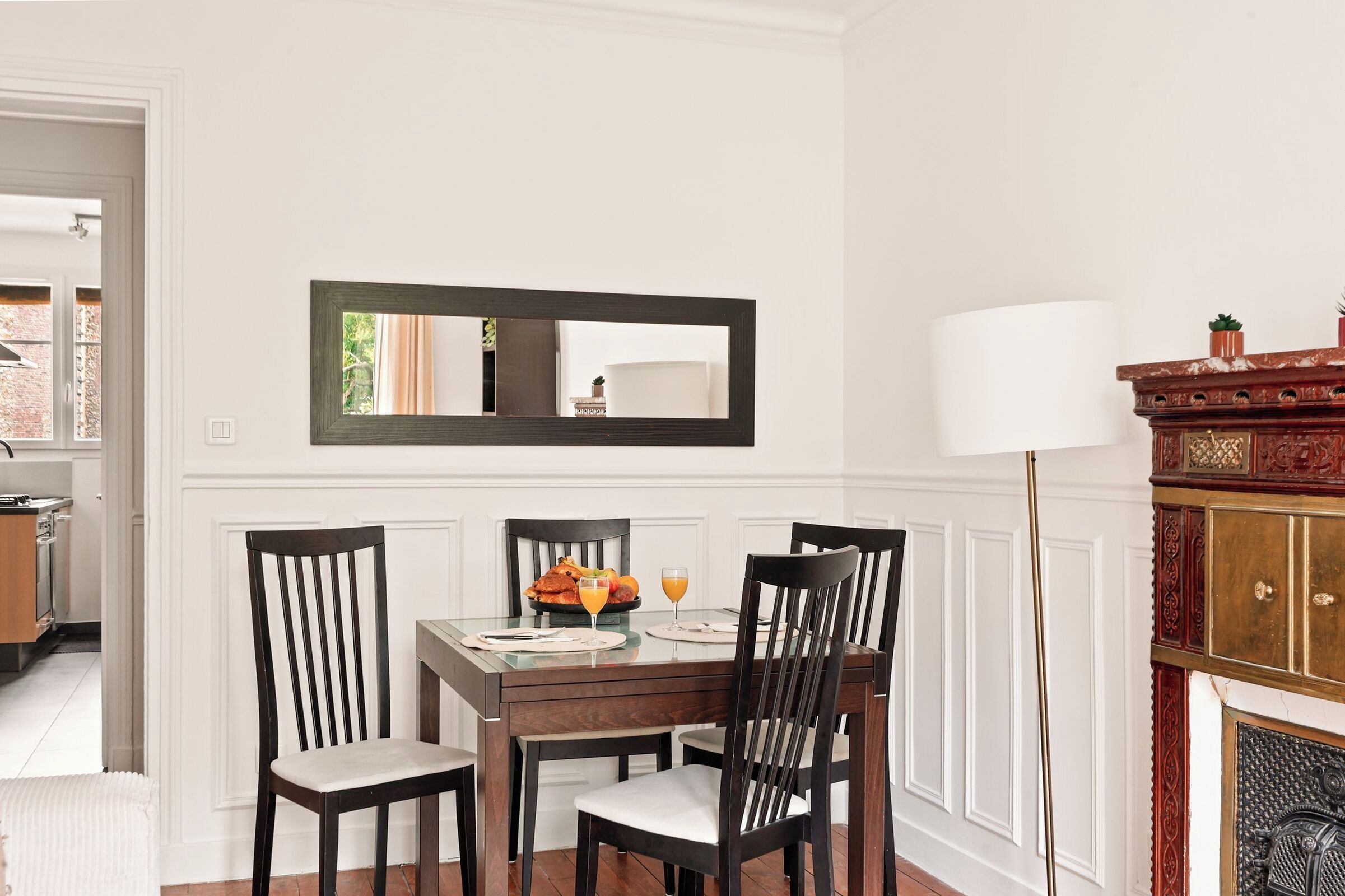 A dining area with a table set for four, a mirror above the table, and a fireplace with a lamp to the right.