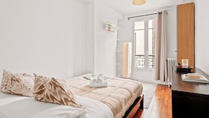 Quarto com cama, mesa de cabeceira, abajur e janela com cortinas.