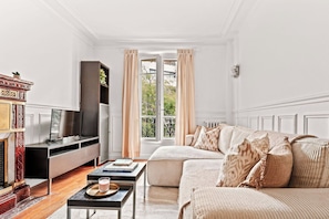 A well-lit living room with a sofa, a television, a fireplace, and a window with curtains. - Elegant Apartment Near Eiffel Tower (Paris)