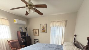 Ein Schlafzimmer mit einem Bett, einem Schreibtisch, einem Stuhl, einem Deckenventilator und einem Fenster mit Vorhängen.