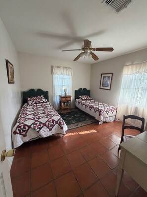 A bedroom with two beds, a ceiling fan, a window with curtains, and a small table with books.