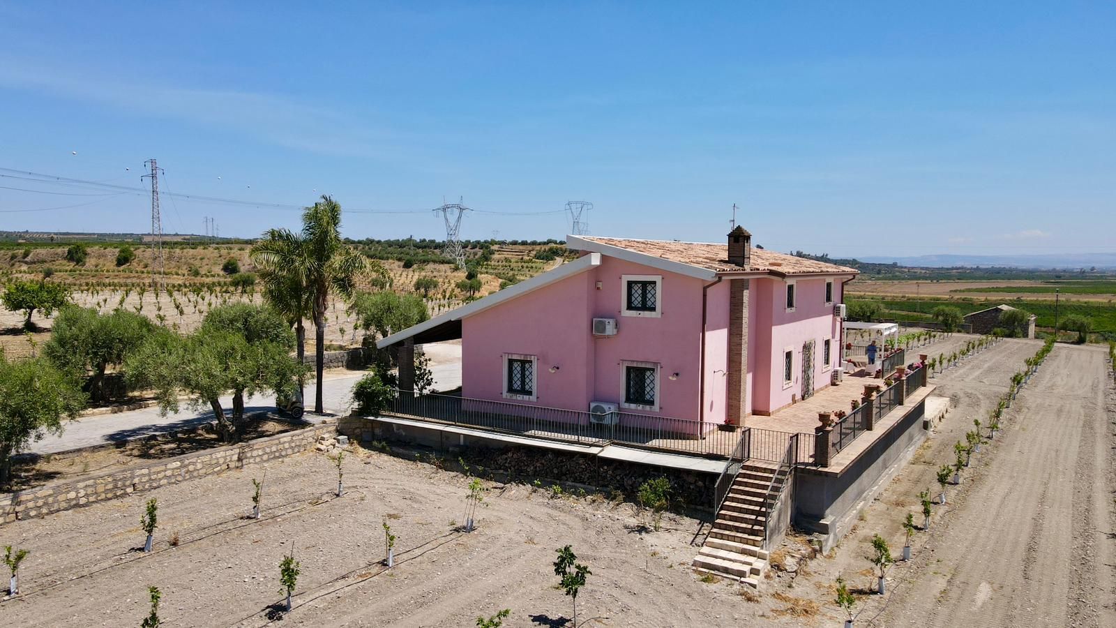 Een roze gebouw met balkon, omgeven door een wijngaard en een onverharde weg.