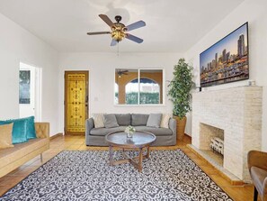 Una sala de estar con un sofá, una mesa de centro, una chimenea, un ventilador de techo y una pintura de un paisaje urbano.