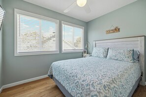 Ein Schlafzimmer mit einem Bett, zwei Fenstern mit Jalousien, einem Nachttisch mit Lampe und einer Wanddekoration.