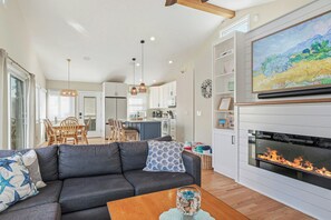 Ein modernes Wohnzimmer mit Kamin, ein Essbereich mit Holztisch und -stühlen sowie eine Küche mit weißen Schränken.