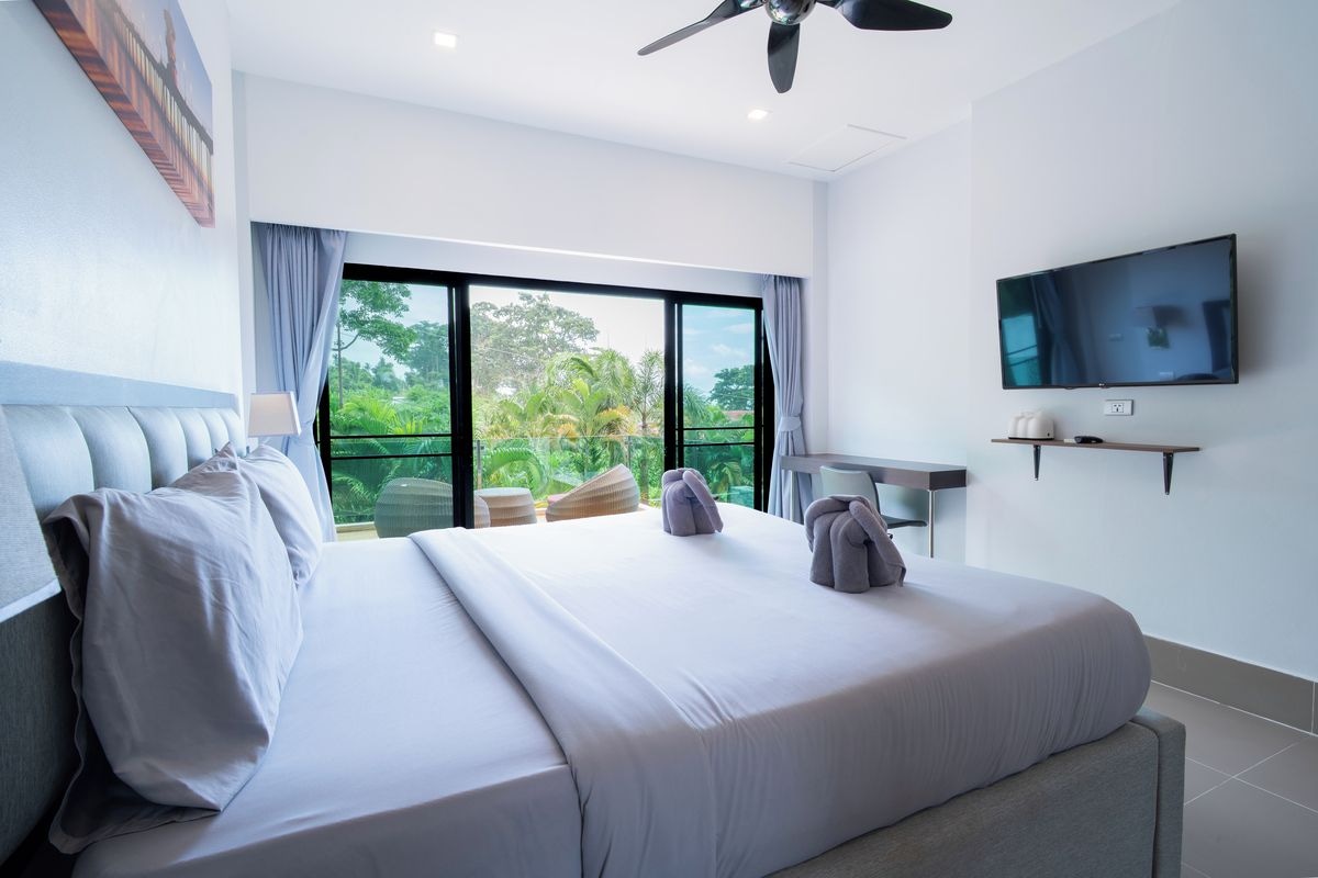 A hotel room with a large bed, a flat-screen TV, a ceiling fan, and a view of greenery outside.