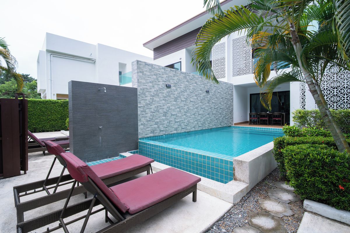 A modern outdoor swimming pool with lounge chairs, a stone wall, and a white building in the background.