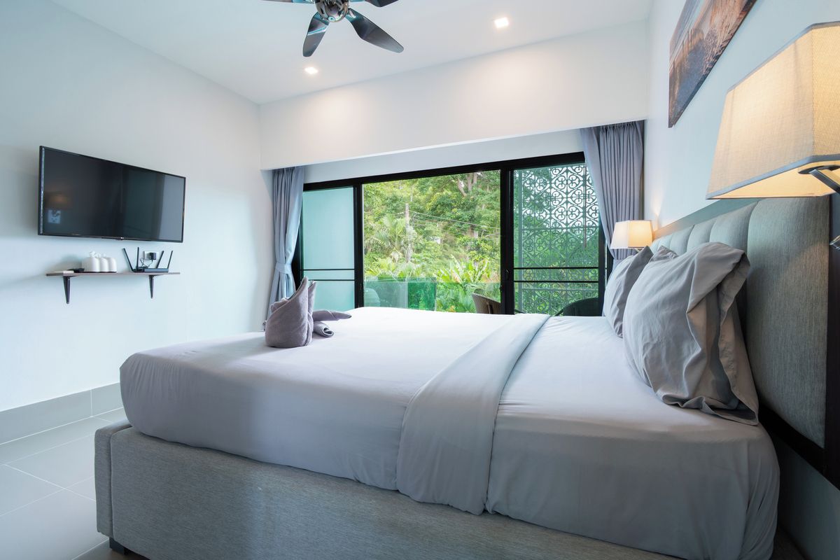 A modern hotel room with a large bed, a flat-screen TV, and a balcony with a view of greenery.