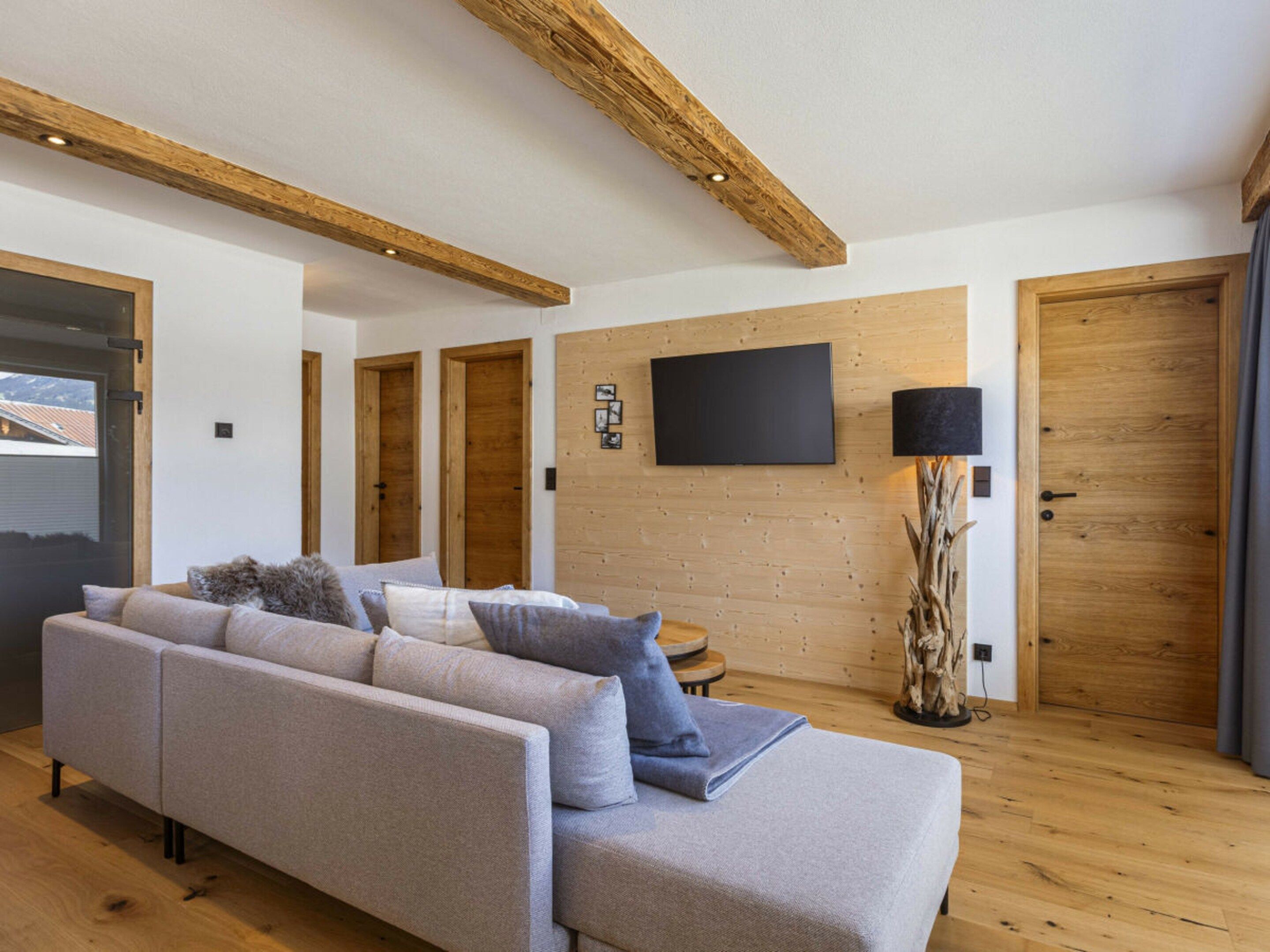 A modern living room with a sofa, a flat-screen TV mounted on the wall, wooden doors, and a large window.