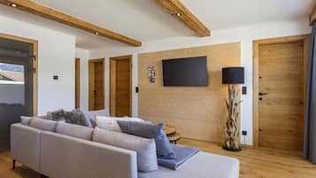A modern living room with a sofa, a flat-screen TV mounted on the wall, wooden doors, and a large window.