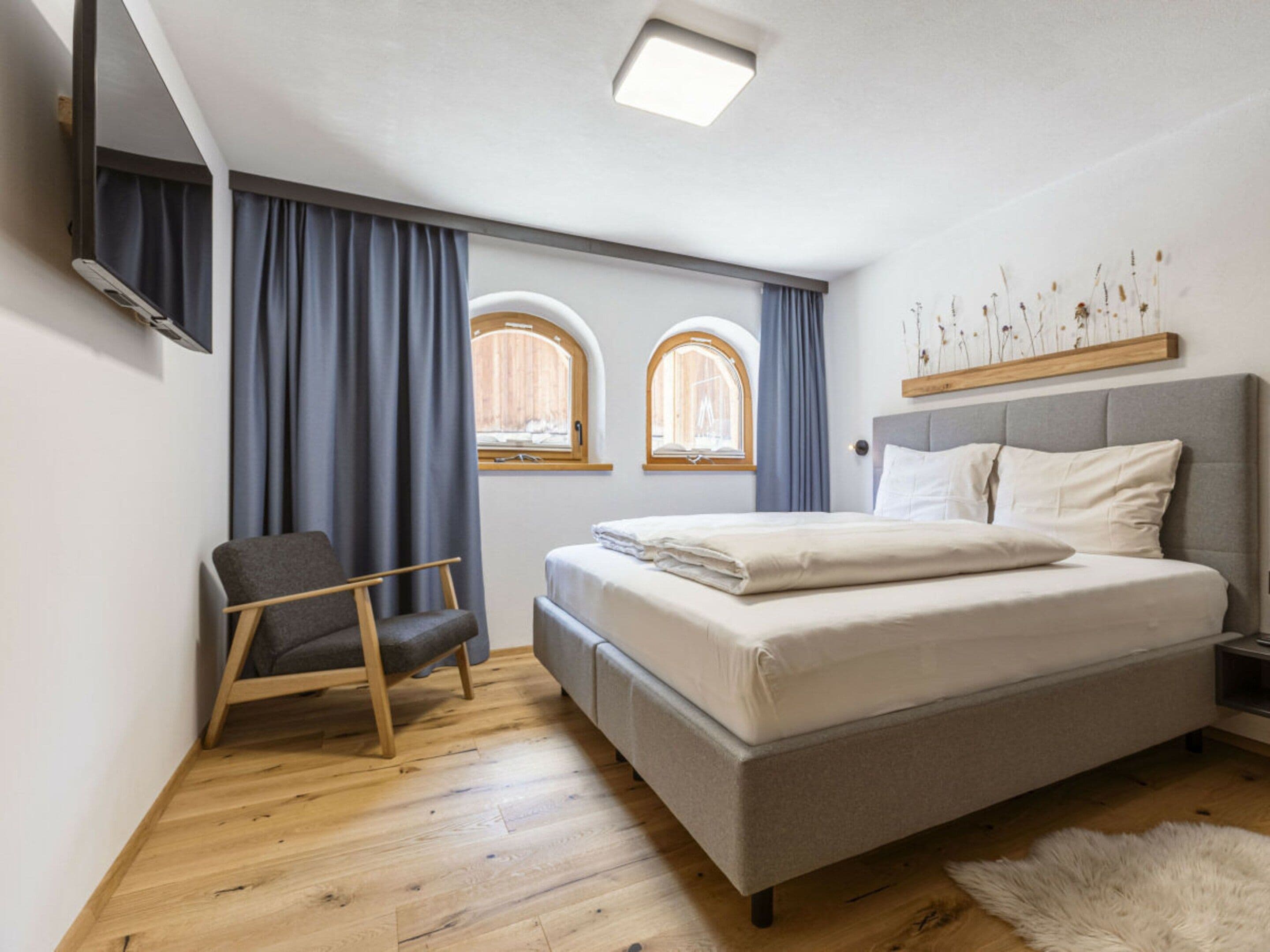 A bedroom with a bed, a chair, two windows, a ceiling light, and a wall-mounted TV.