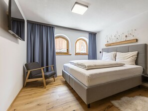 Un dormitorio con una cama, una silla, dos ventanas, una lámpara en el techo y un televisor montado en la pared.