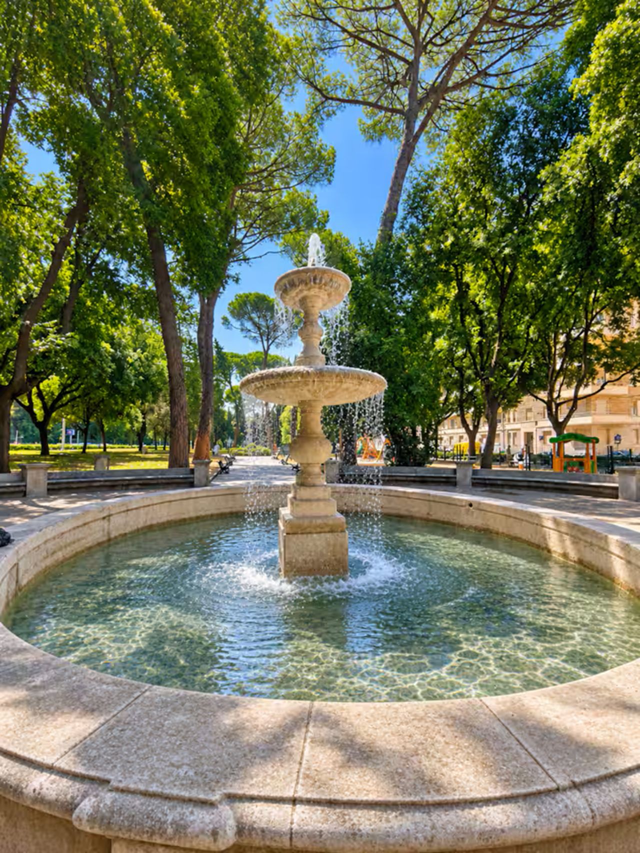 A multi-tiered fountain in a circular stone basin surrounded by trees.