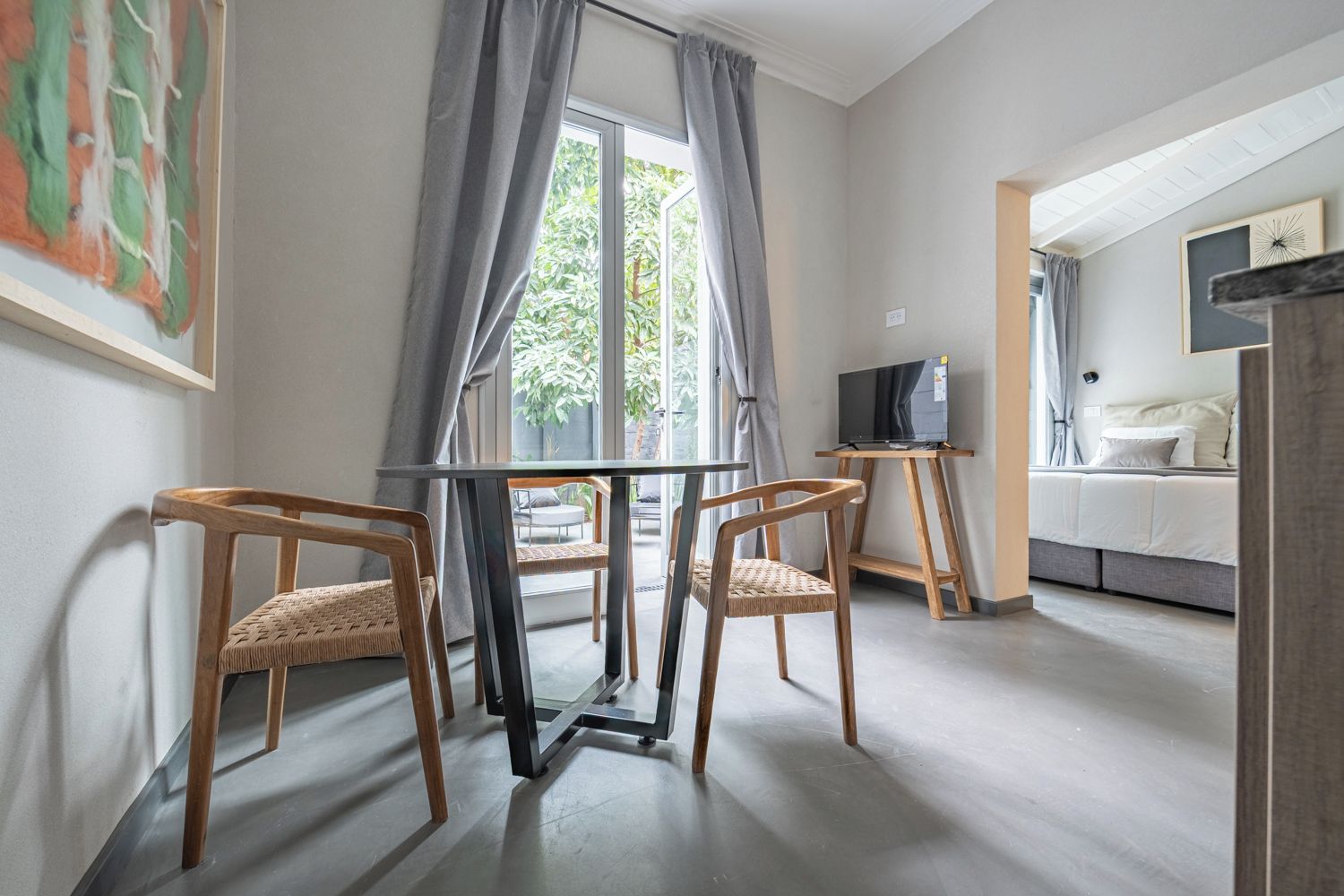 Una habitación con una mesa, sillas, un televisor sobre un mueble bajo y una cama con cabecera.