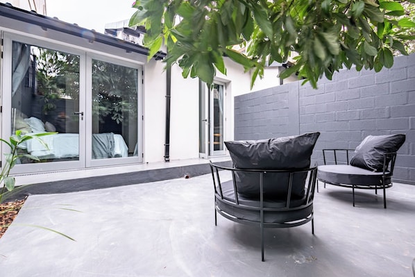 A modern outdoor seating area with two chairs and a table, a glass door leading to a room, and a grey brick wall.