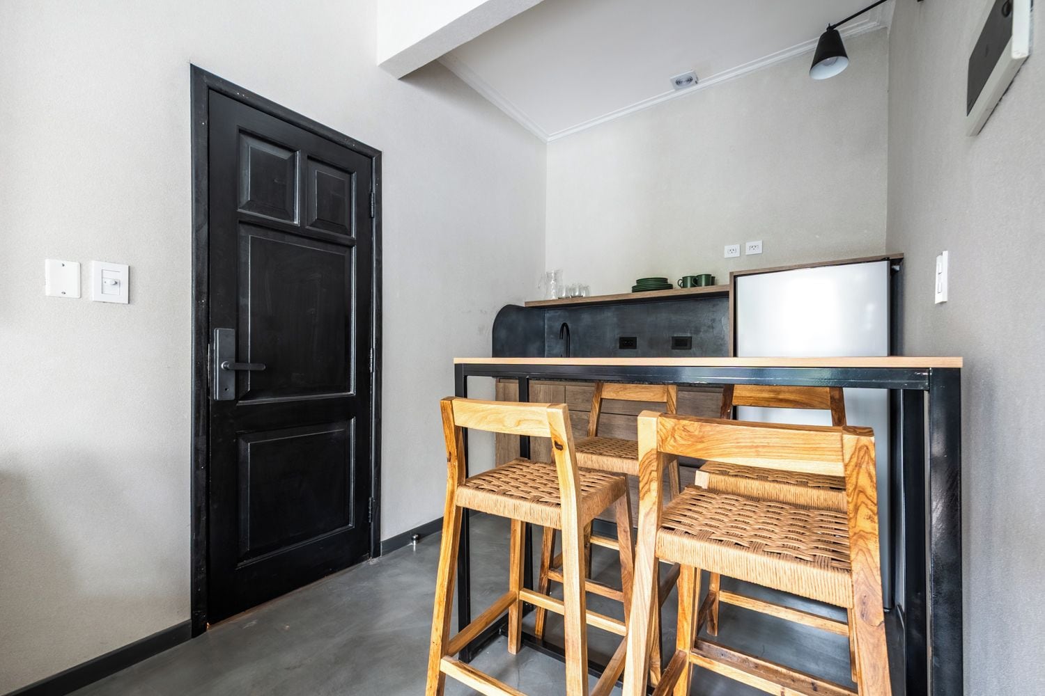 Cocina compacta con puerta negra, un pequeño comedor con dos sillas de madera y un refrigerador blanco.