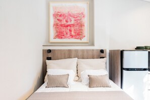 A neatly made bed with a headboard, a framed abstract painting above it, and a refrigerator to the right.