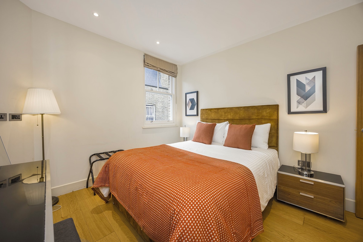 A hotel room with a bed, bedside tables, a lamp, a window, and artwork on the wall.