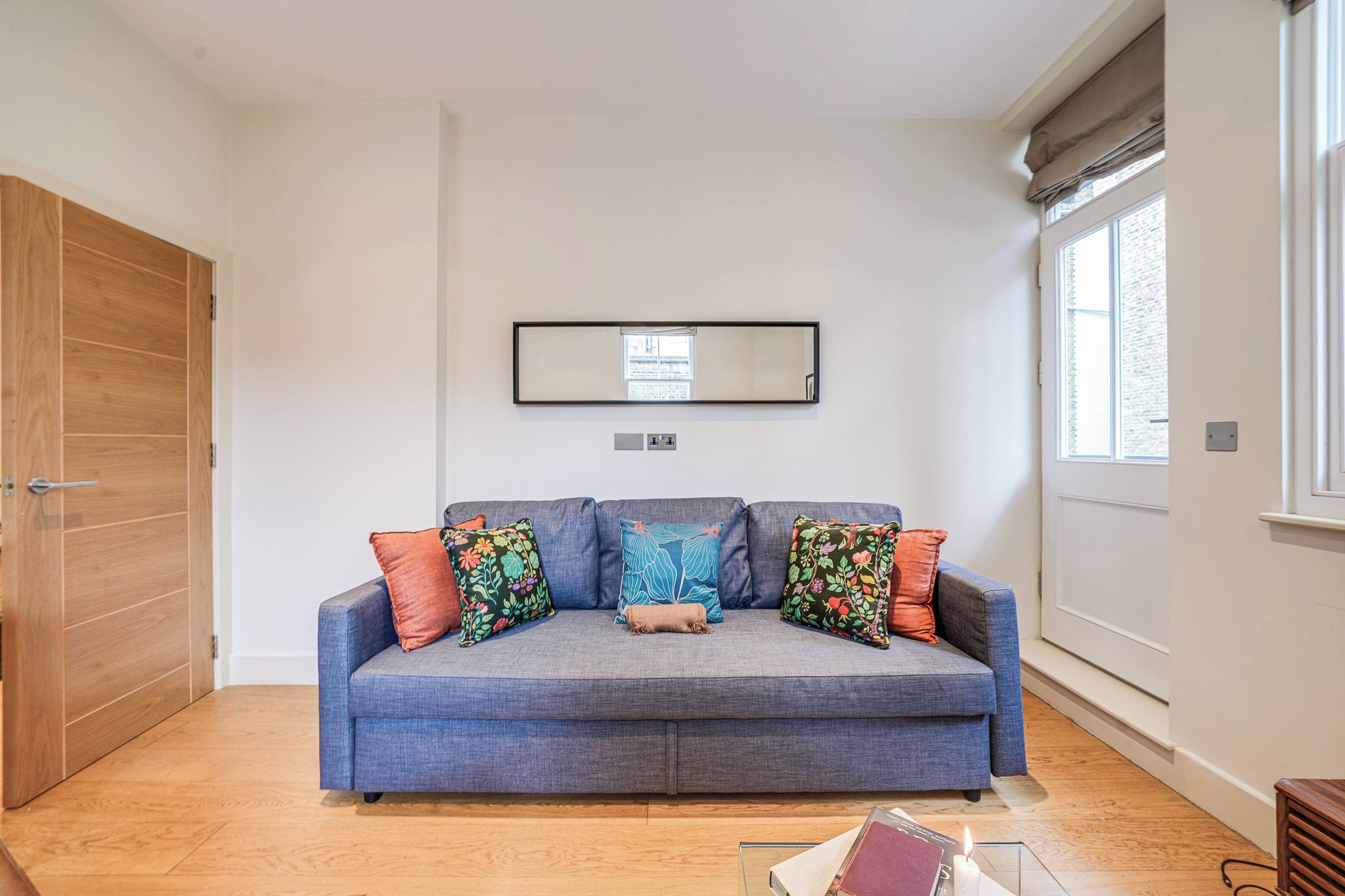 Ein Zimmer mit einem blauen Sofa, zwei blumigen Kissen, einem kleinen beigen Kissen, einem gerahmten Spiegel, einer Holztür und einem Fenster mit Blick.