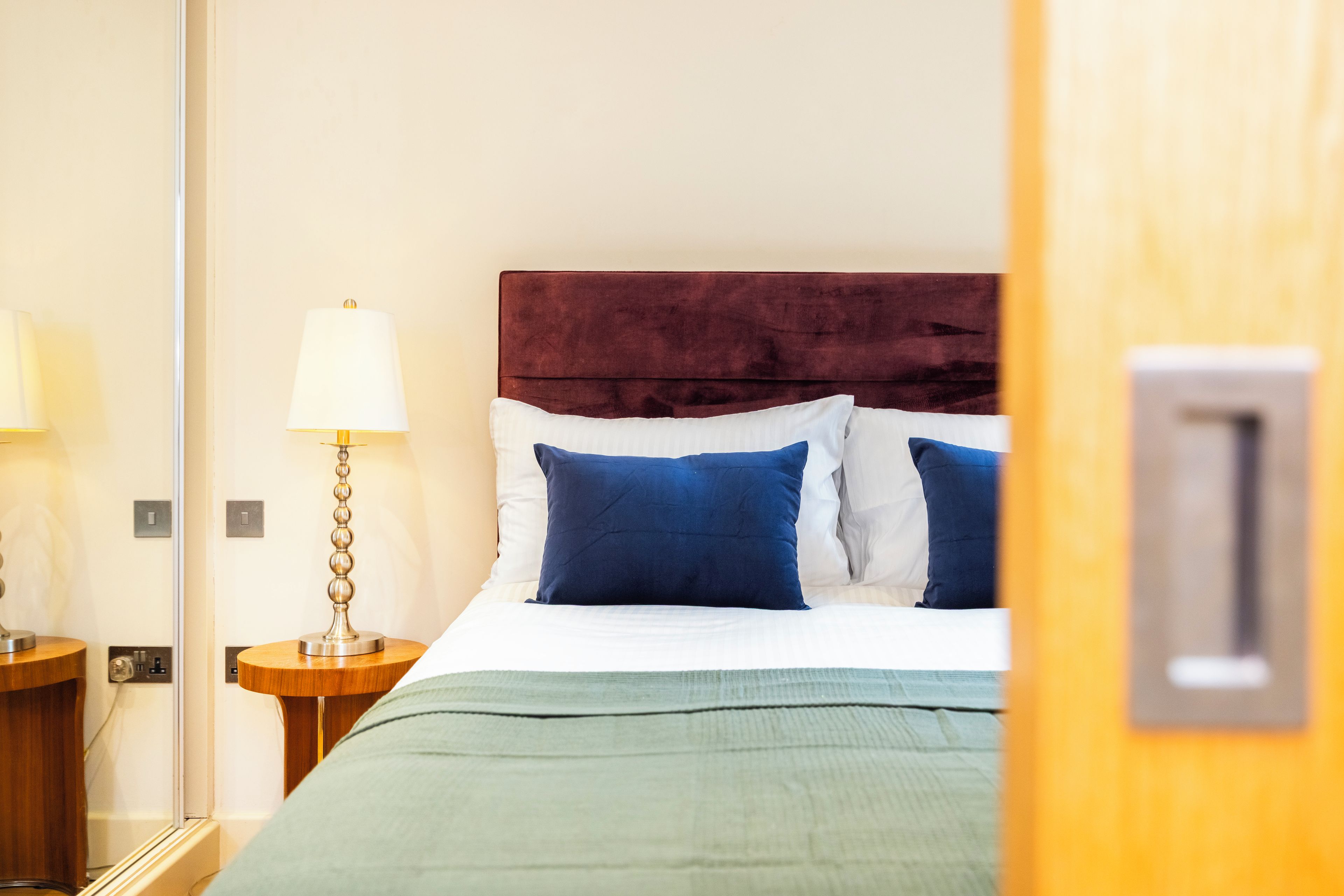 Una habitación de hotel con una cama, una mesita de noche, una lámpara y una puerta con manija.