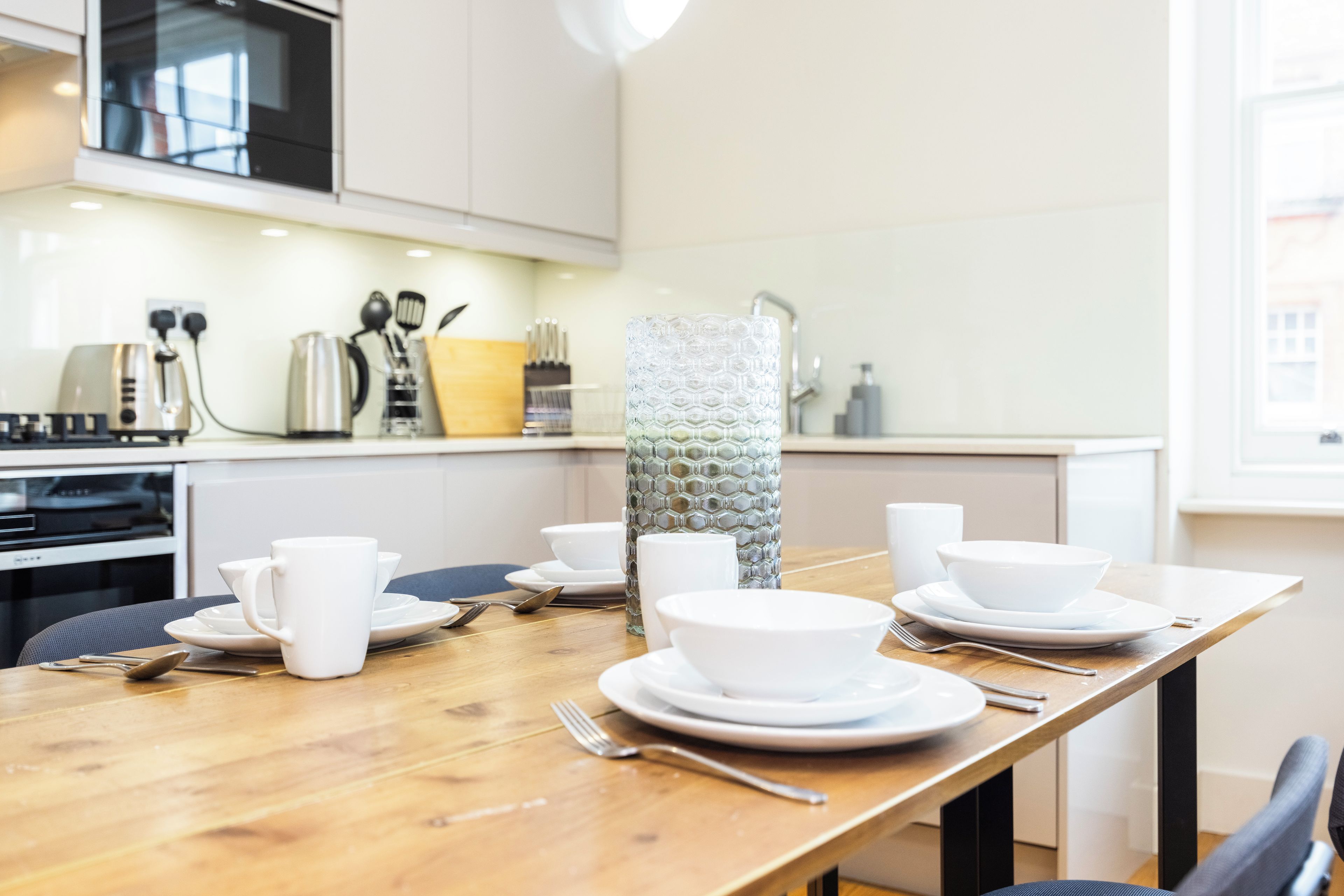 Een moderne keuken met witte kasten, een houten eettafel gedekt met witte borden, kommen en kopjes.