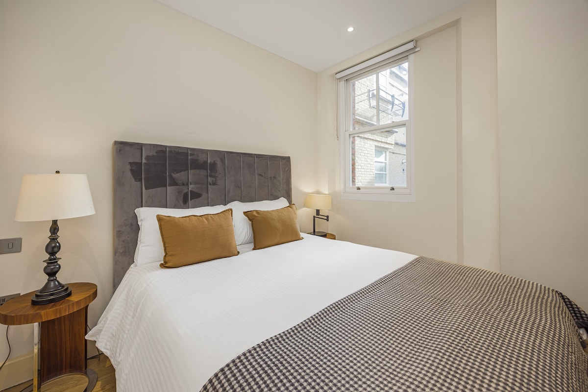 A hotel room with a large bed, two bedside lamps, a window with a view of buildings, and a wooden nightstand.