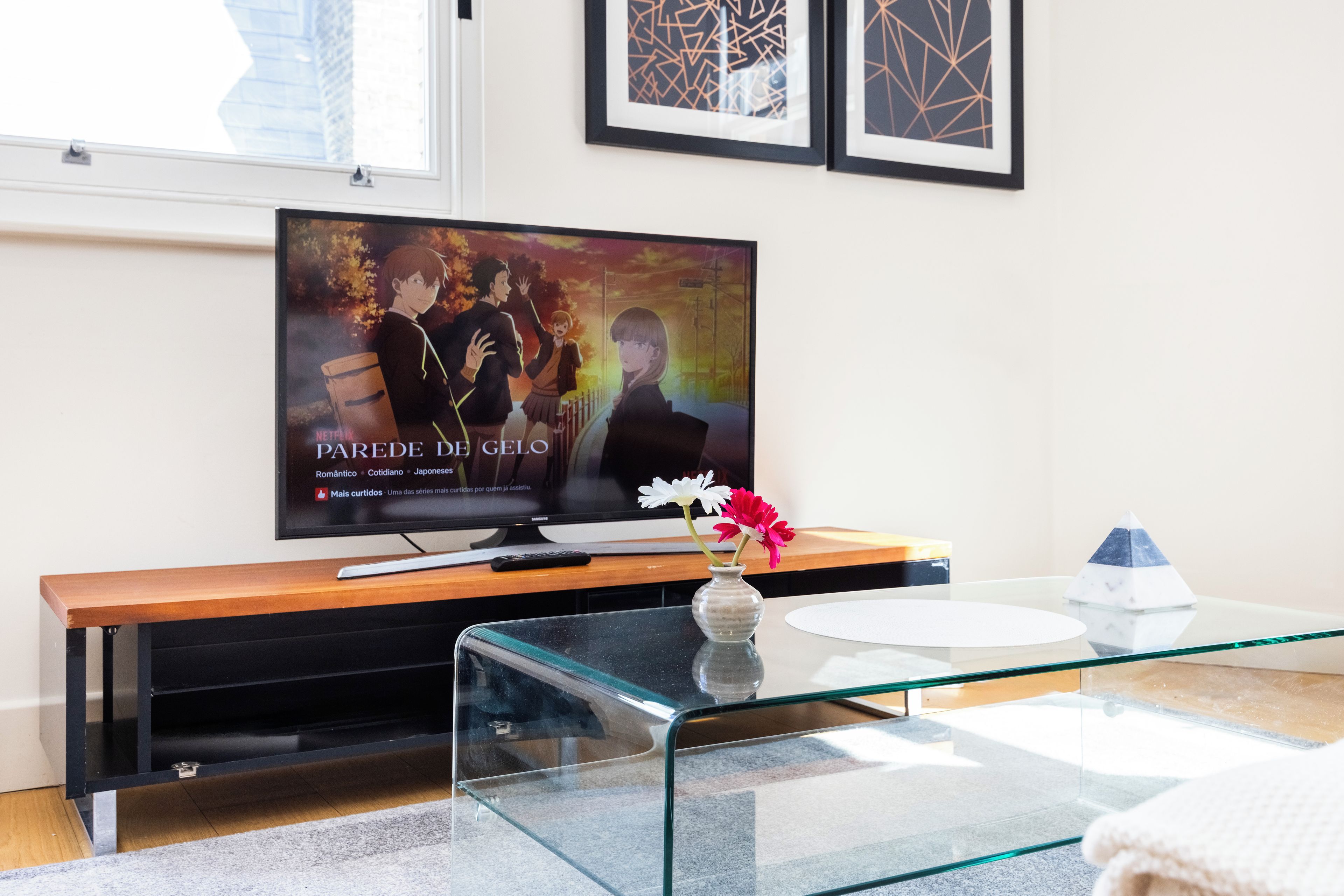 Een moderne woonkamer met een flatscreen televisie op een houten standaard, een glazen salontafel en een vaas met bloemen.