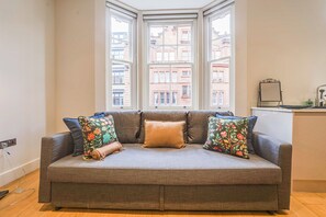 A grey sofa with floral cushions in a room with a bay window overlooking a cityscape.
