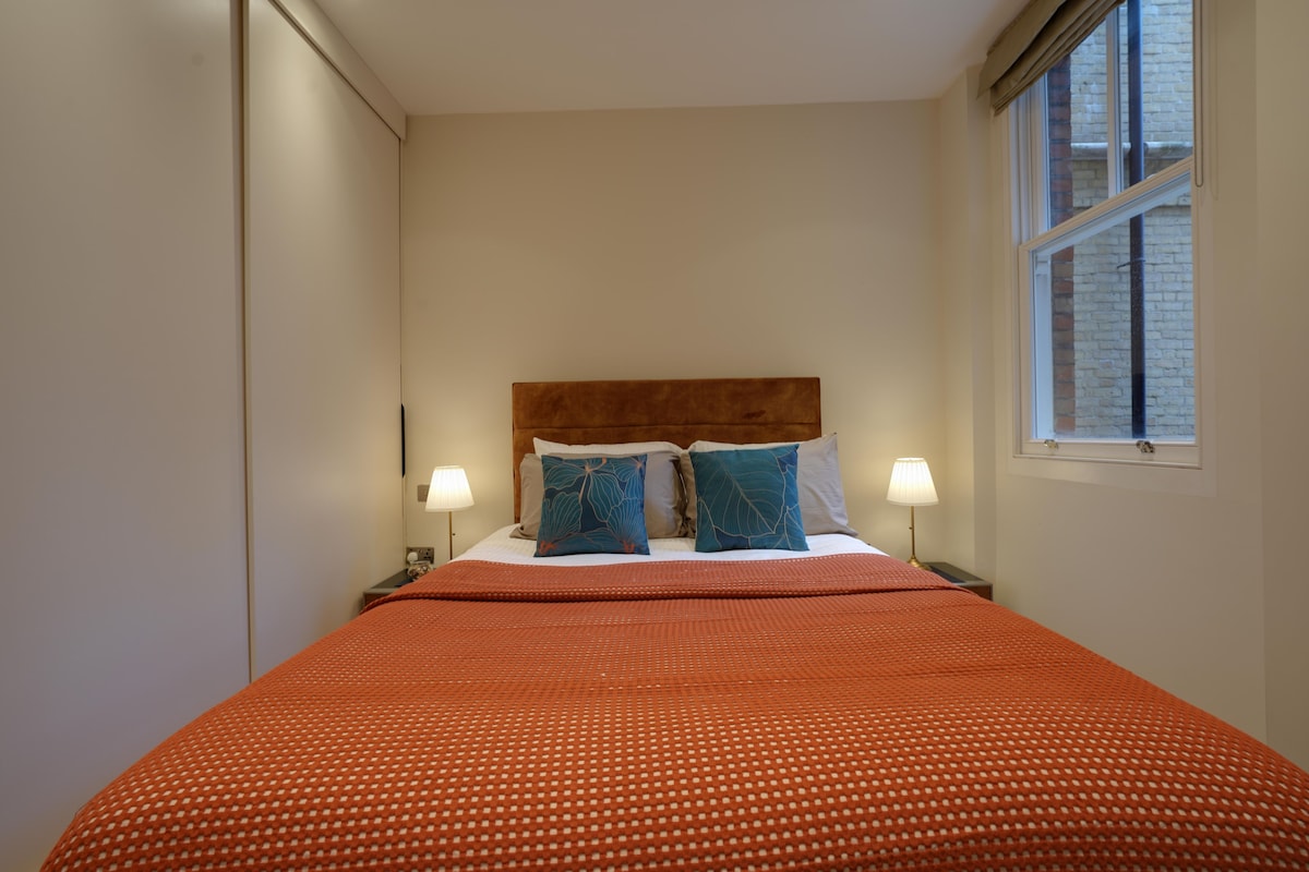 A neatly made bed with a patterned bedspread, two pillows with a leaf design, and a wooden headboard. There is a window with a view of a brick building.