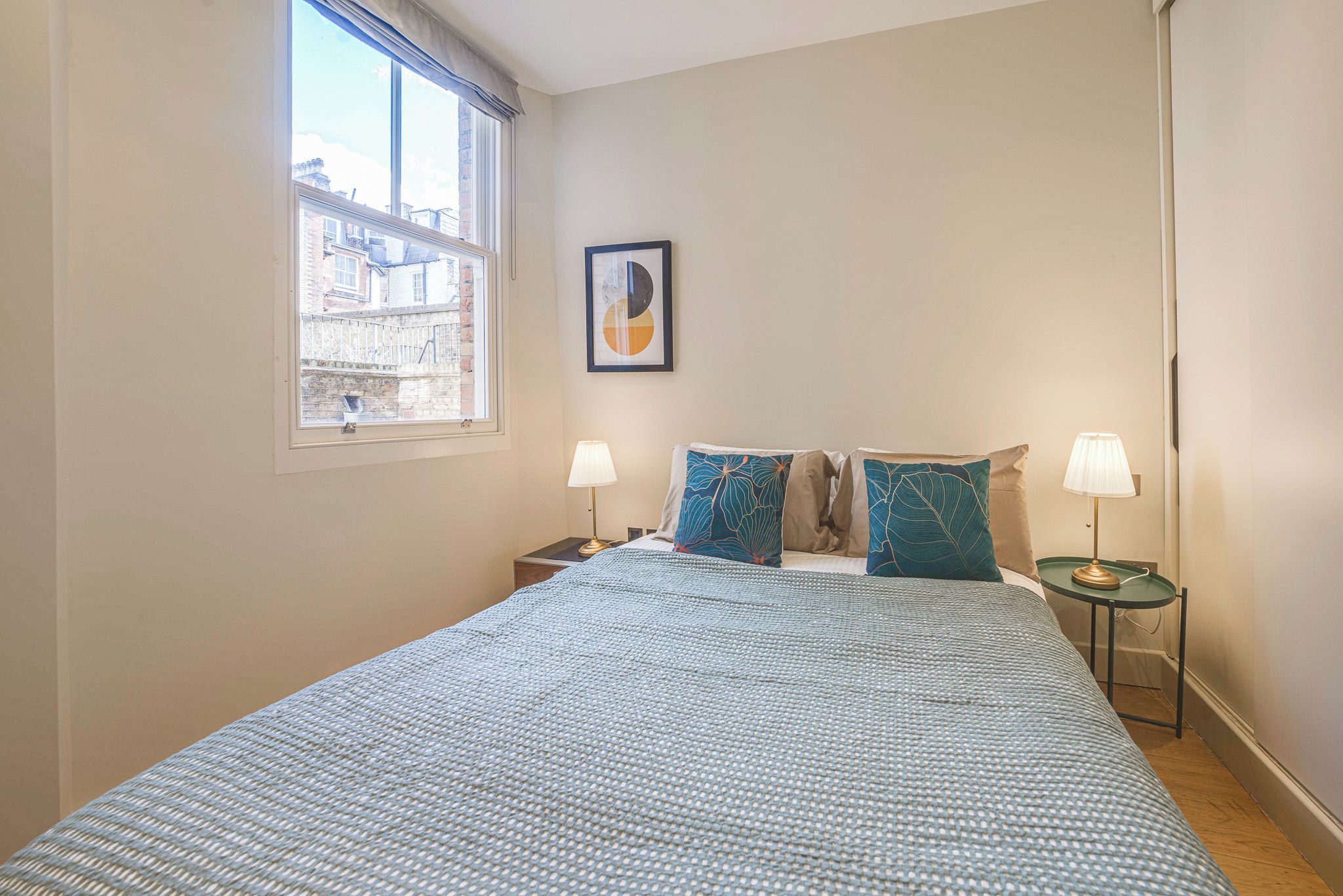 A bedroom with a large bed, two bedside tables with lamps, a window with a view of buildings, and a framed picture on the wall.