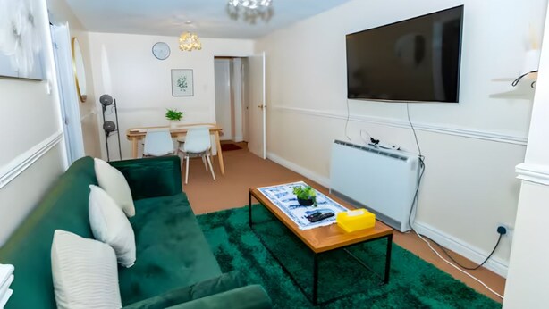 A living room with a green sofa, a coffee table, and a TV mounted on the wall. - Cozy 1BR Ground Floor Maisonette in Town Centre (Bletchley)