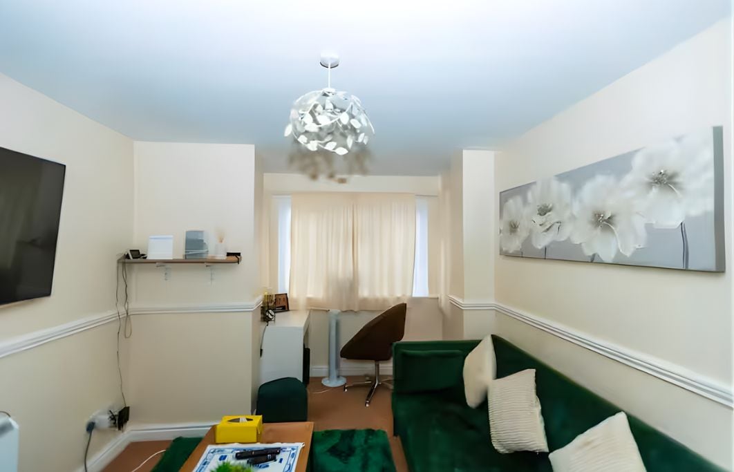 Un salon avec un canapé vert, un téléviseur à écran plat, un luminaire au plafond et un grand tableau représentant des motifs floraux accroché au mur.
