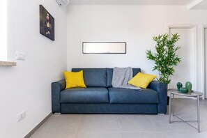 Una sala de estar moderna con un sofá azul oscuro, dos almohadas amarillas, una manta gris, una obra de arte enmarcada en la pared, una planta de bambú en maceta y una mesita con objetos decorativos.