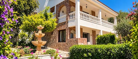A two-story house with a stone facade, a balcony with a balustrade, and a garden with a fountain.