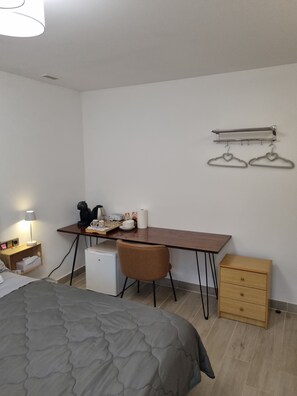 Una camera compatta con un letto, una scrivania, una sedia, un piccolo frigorifero, un comodino con una lampada e un tavolino in legno.