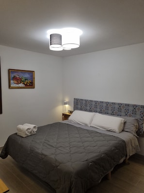 Una cama bien hecha con colcha gris, cabecera con diseño estampado, un cuadro de frutas en la pared y una lámpara de noche.