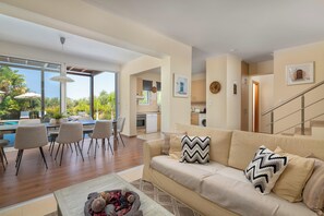 A modern living room with a beige sofa, a dining area with chairs, a kitchen in the background, and a view of a swimming pool. - Villa Olvia - Three Bedroom Villa, Sleeps 6 (Rhodes)