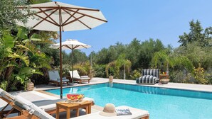Zona junto a la piscina con sillones, sombrillas y una mesa con frutas y un sombrero.