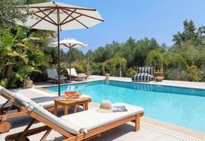 A poolside area with lounge chairs, umbrellas, and a table with fruits and a hat. - Villa Olvia - Three Bedroom Villa, Sleeps 6 (Rhodes)