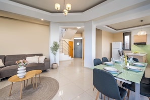 A modern living room with a sofa, coffee table, dining table set for four, and a staircase leading to another level. - Villa Aqua - Three Bedroom Villa, Sleeps 7 (Lachania)