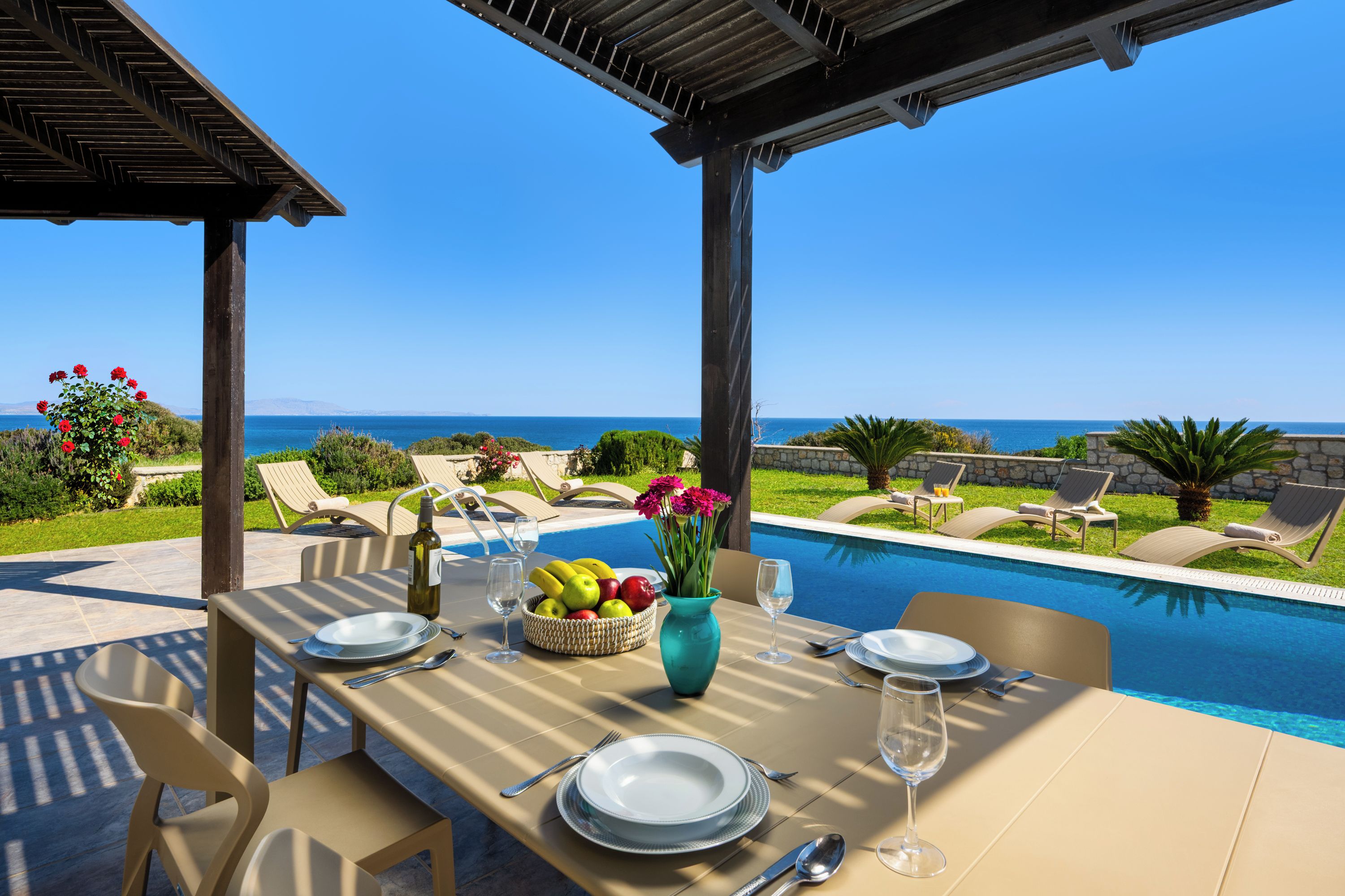 Un espace repas aménagé au bord de la piscine, avec une table dressée pour deux personnes, un vase de fleurs et un saladier de fruits.