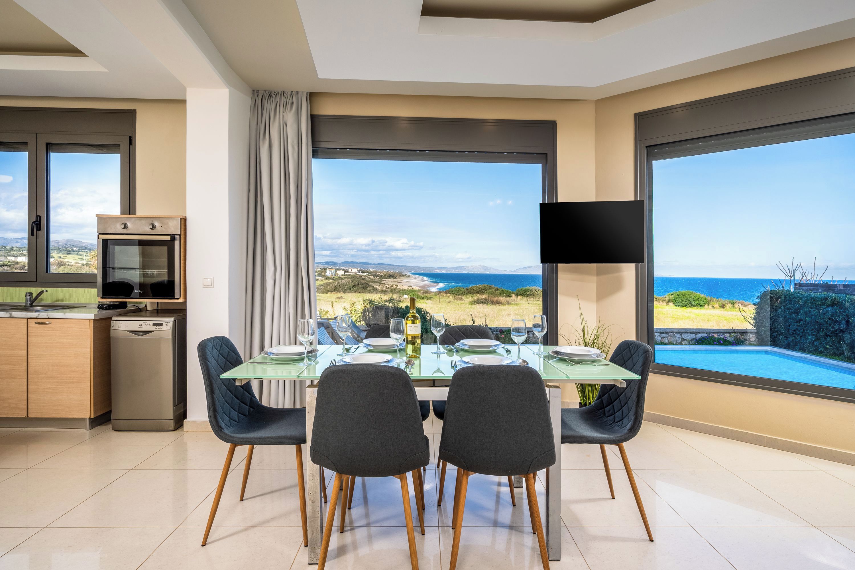 Ein moderner Essbereich mit Glastisch, zwei Stühlen, einer Küche mit Geschirrspüler und einem großen Fenster mit Meerblick.