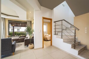 A modern living room with a sofa, a coffee table, and a potted plant. There is a staircase with a metal railing leading to a bedroom. - Villa Aqua - Three Bedroom Villa, Sleeps 7 (Lachania)