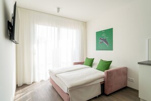 A hotel room with a bed, a TV mounted on the wall, a window with sheer curtains, and a green wall art piece. - eeeden apartments Wien 10 (Wien)